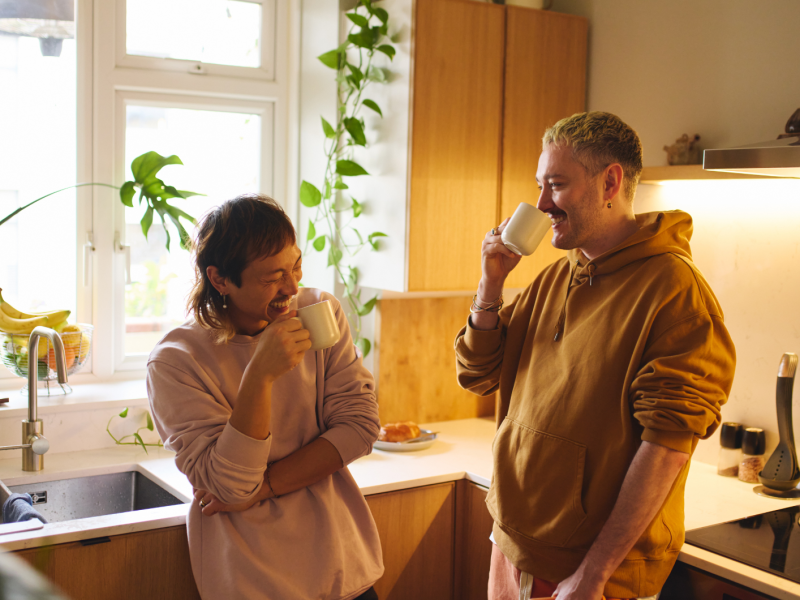 Frau und Mann in der Küche, die beide an einer Tasse nippen und lachen.