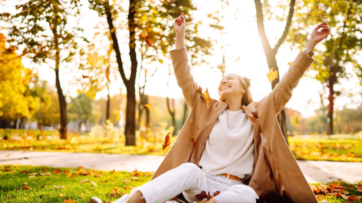 Frau im Park auf dem Rasen, die herbstliche Blätter in die Luft wirft und lacht