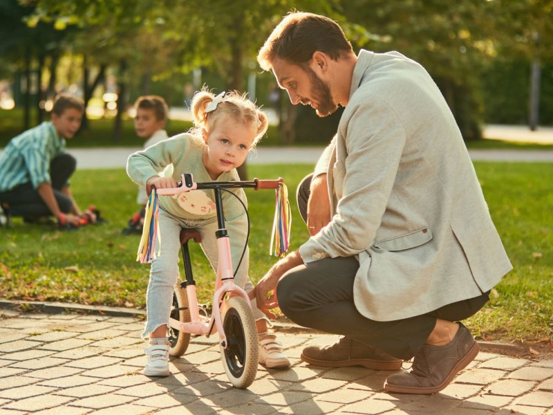 Vater kniet draußen im Park neben seiner Tochter, die auf einem Laufrad sitzt.