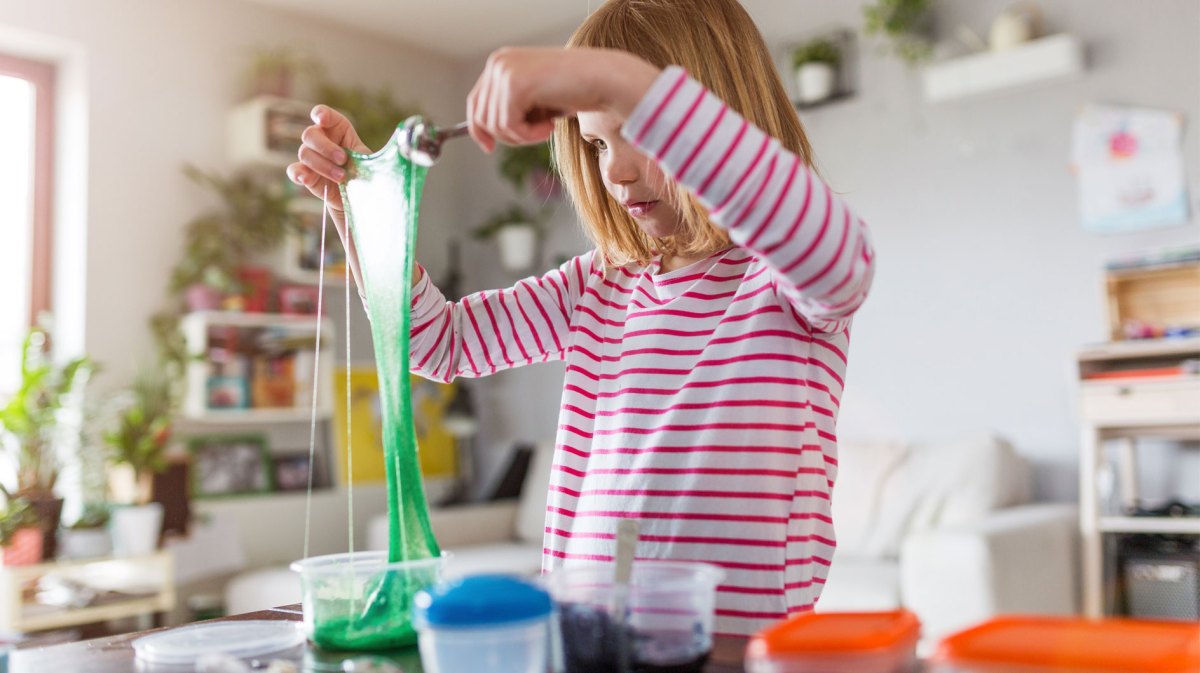 Mädchen mit blonden Haaren zieht grünen Schleim in die Höhe, umgeben von Spielzeug und Pflanzen.