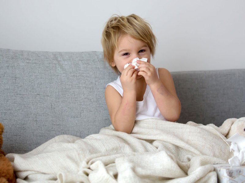 Junge, ca. 4 Jahre, sitzt im Unterhemd und zugedeckt auf der Couch und schnäuzt sich die Nase.