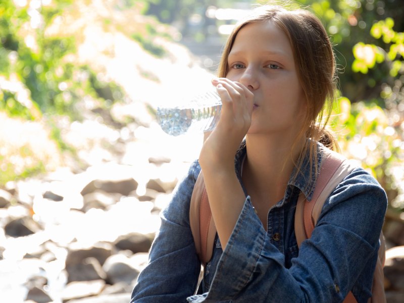 Mädchen sitzt an einem Bach im Wald und trinkt einen Schluck Wasser aus der Flasche.