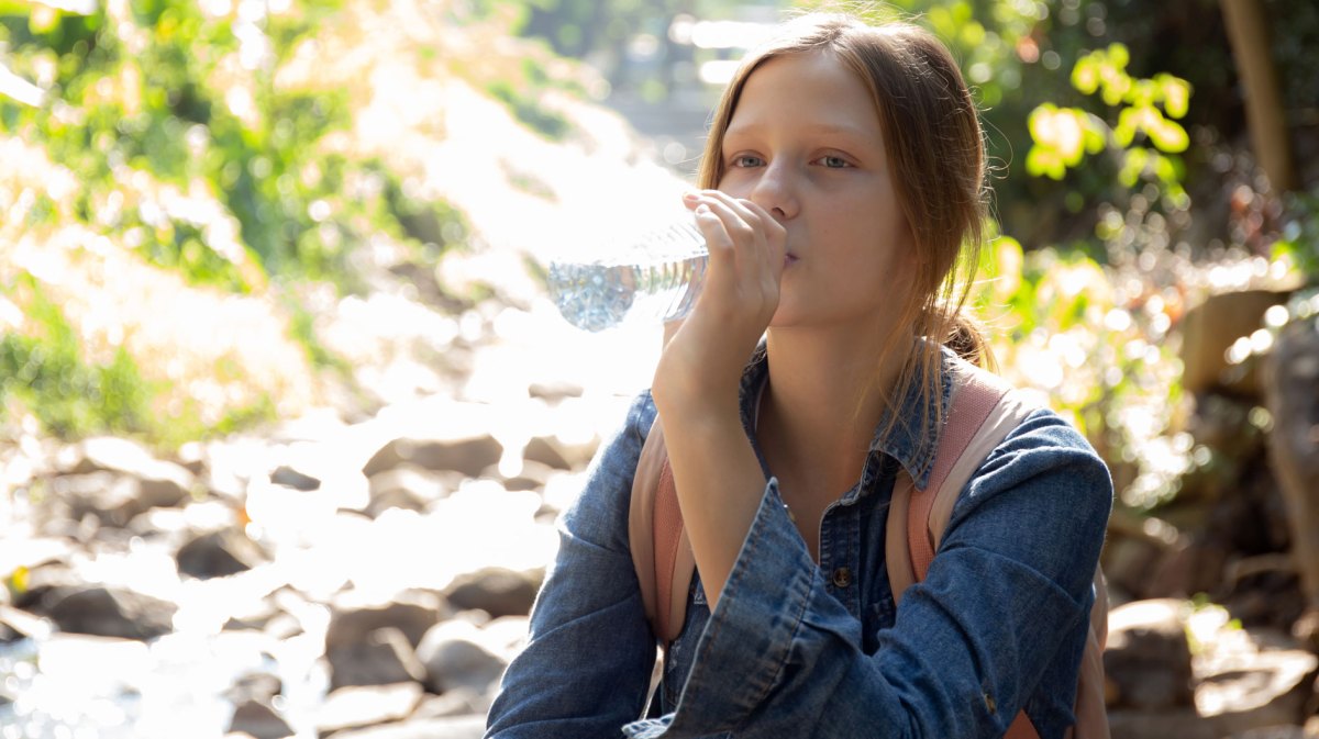 Mädchen sitzt an einem Bach im Wald und trinkt einen Schluck Wasser aus der Flasche.