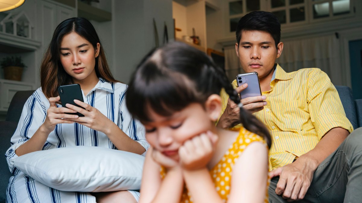 Eltern sitzen mit ihren Smartphones auf dem Sofa, die Tochter hockt gelangweilt im Vordergrund und weiß nichts mit sich anzufangen.