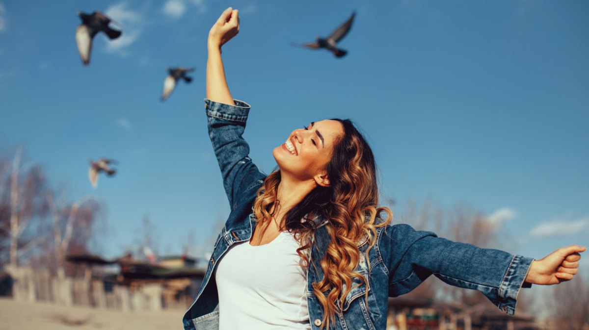 Frau mit Locken streckt die Arme in die Luft. Im Hintergrund sieht man blauen Himmel und Tauben