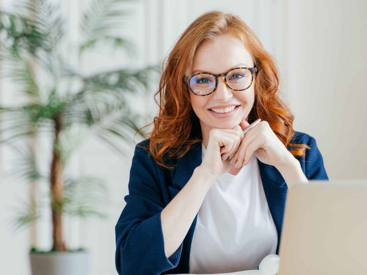 Frau mit roten Haaren und Brille sitzt am Schreibtisch.