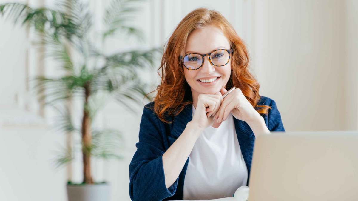 Frau mit roten Haaren und Brille sitzt am Schreibtisch.