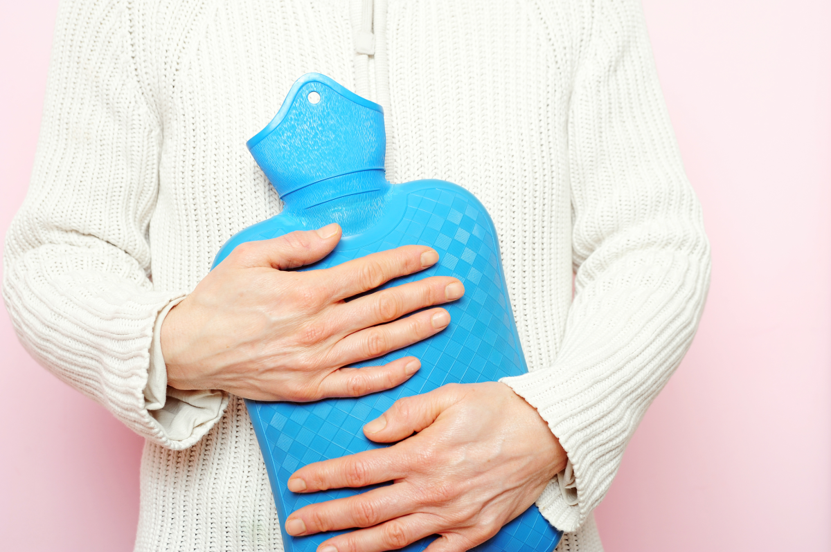 Hände umklammern eine blaue Wärmflasche vor weißem Strickpullover auf rosa Hintergrund.