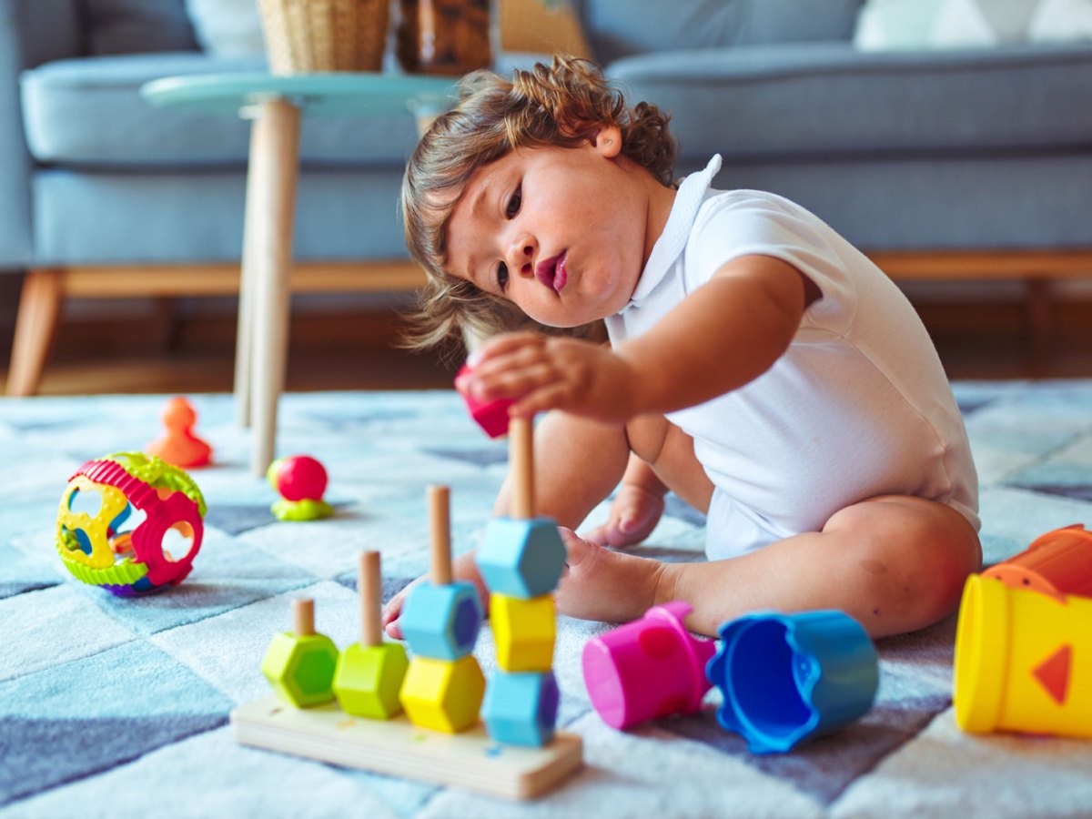 Kind, ca. zwei Jahre, sitzt auf dem Fußboden des Wohnzimmers und spielt konzentriert mit Stapelspielzeug.