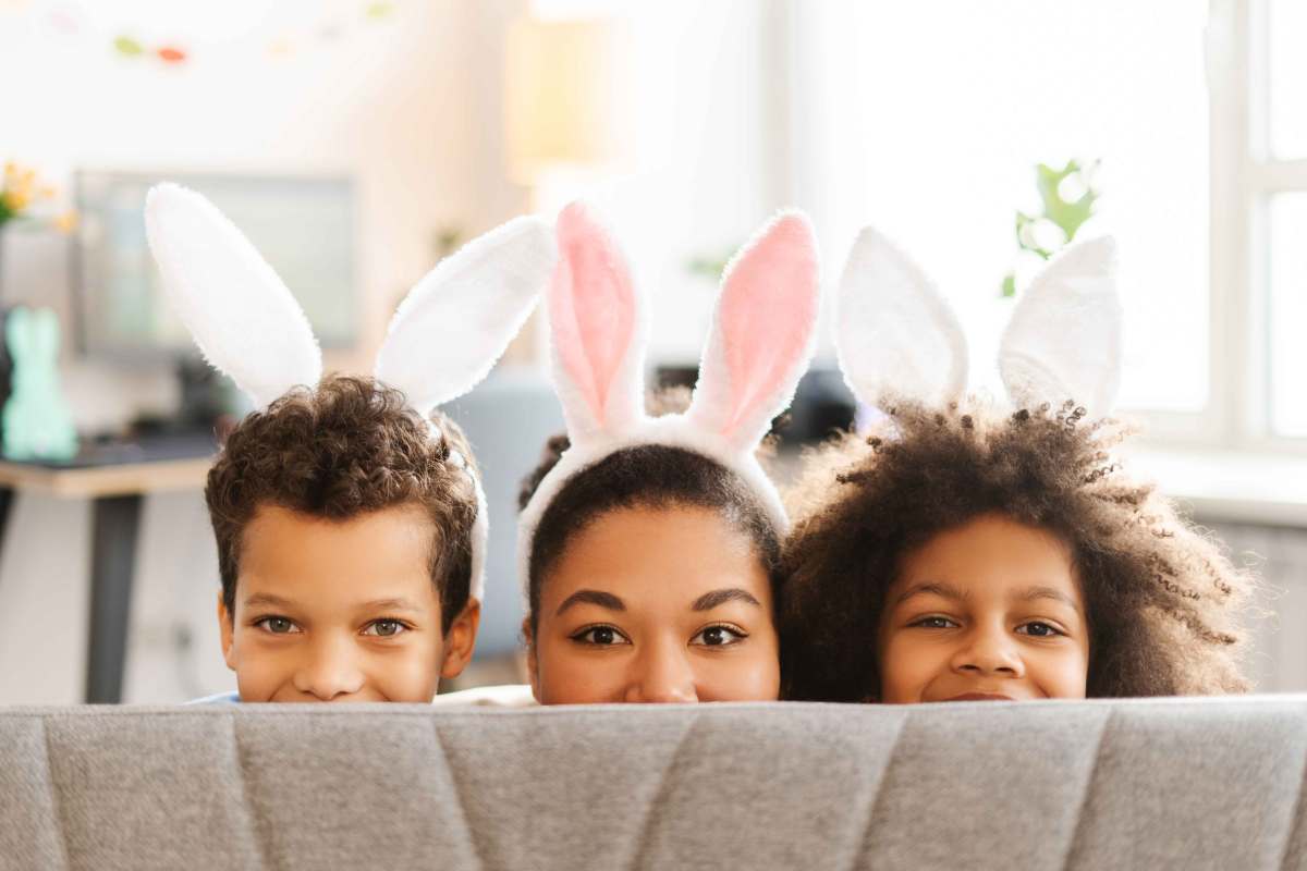 Mutter und zwei Kinder schauen mit Hasenohren auf dem Kopf verschmitzt über die Sofalehne.