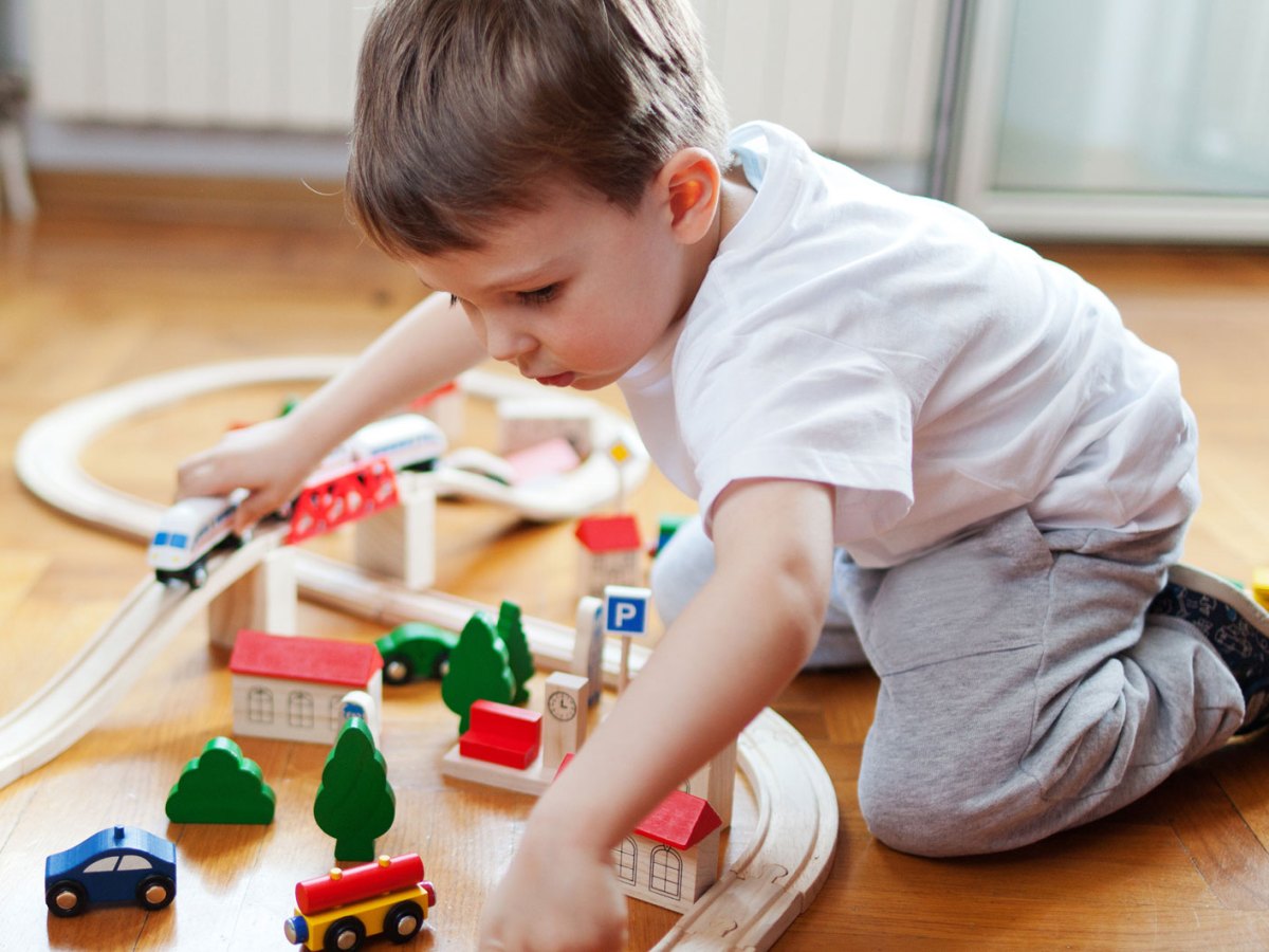 Junge, ca. 3 Jahre, spielt auf dem Boden mit einem Holzzug, den er auf Holzschienen fahren lässt.