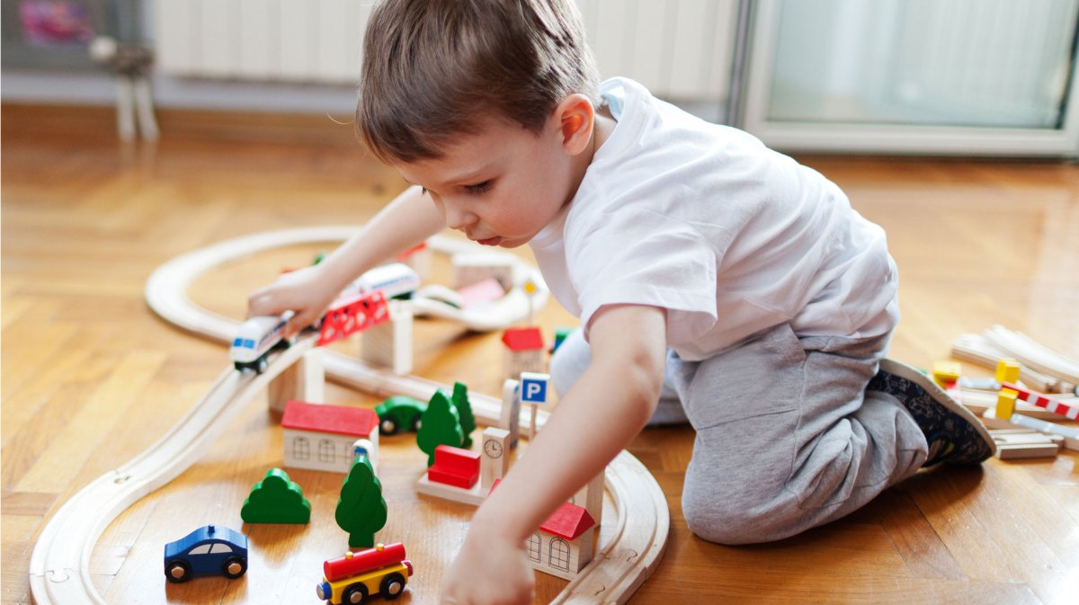 Junge, ca. 3 Jahre, spielt auf dem Boden mit einem Holzzug, den er auf Holzschienen fahren lässt.