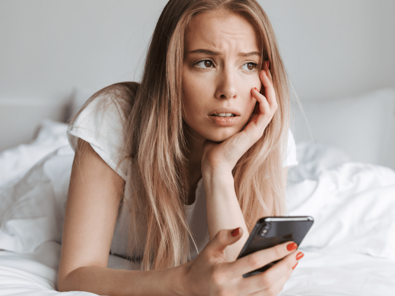 Eine junge Frau mit langen blonden Haaren liegt nachdenklich auf einem Bett, stützt ihren Kopf und hält ein Smartphone in der Hand.