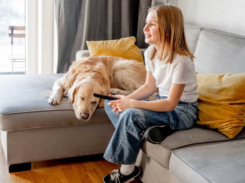 Mädchen, ca 11 oder 12 Jahre alt, sitzt neben ihrem Hund auf dem Sofa, die Fernbedienung in der Hand.
