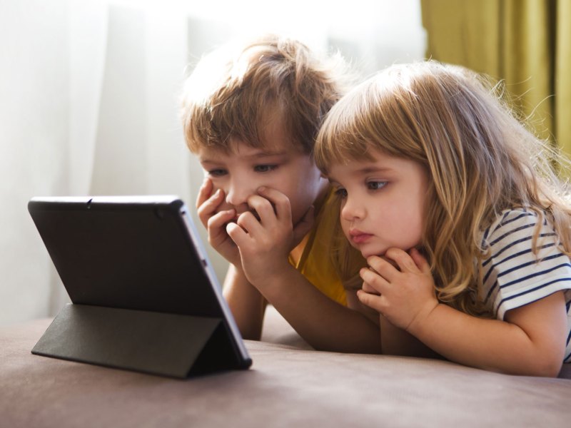 Junge und Mädchen liegen auf dem Boden und schauen etwas auf einem Tablet, das unmittelbar vor ihnen steht.