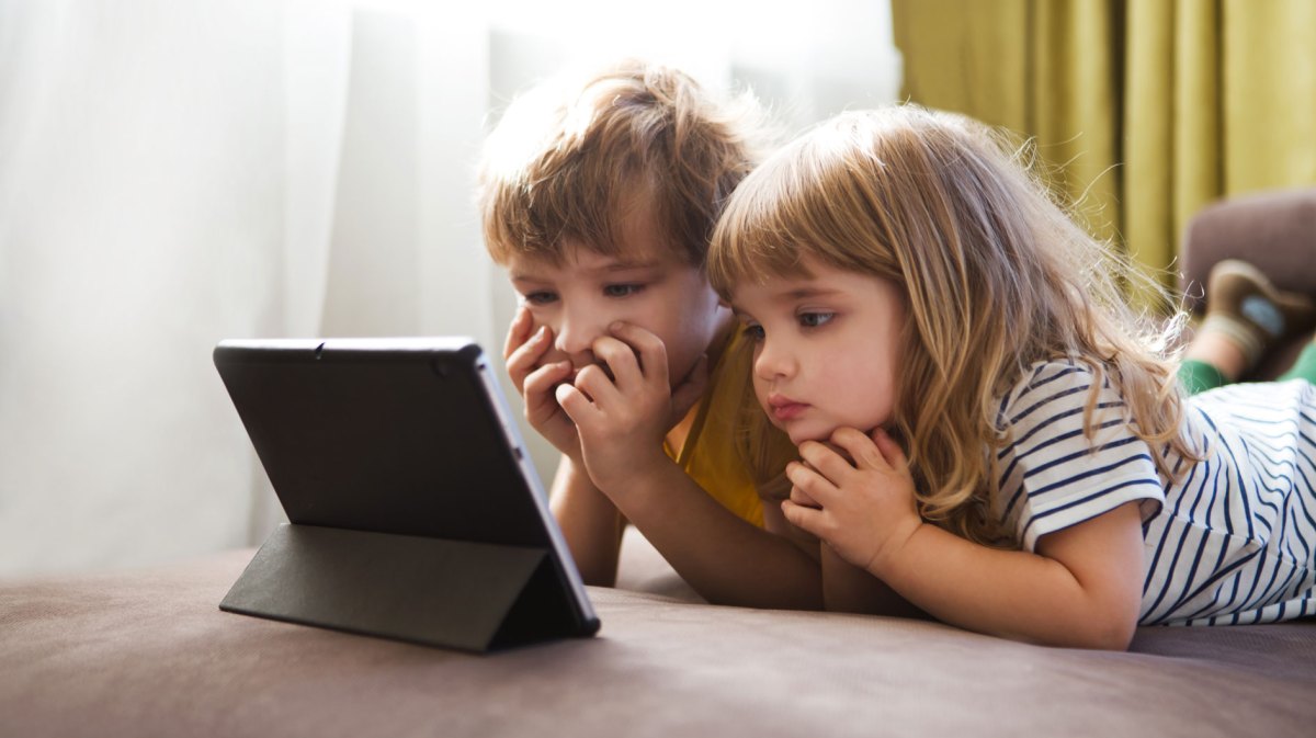 Junge und Mädchen liegen auf dem Boden und schauen etwas auf einem Tablet, das unmittelbar vor ihnen steht.