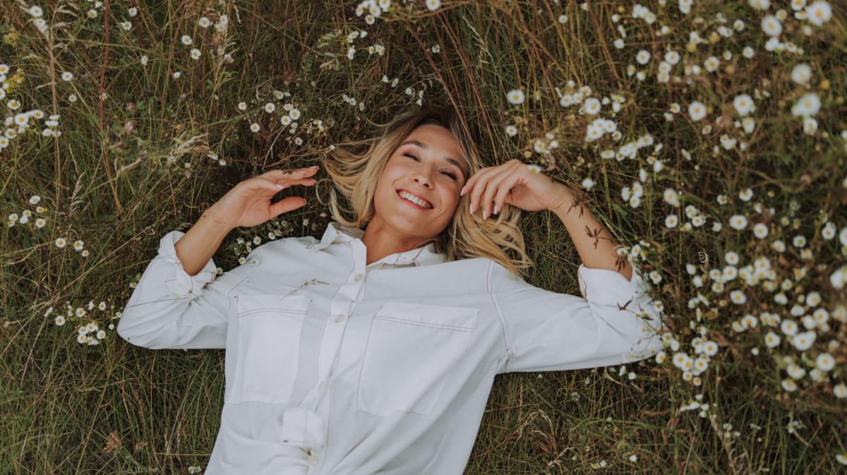 Frau auf einem Rasen mit Blumen, die eine weiße Bluse anhat und in die Kamera lächelt