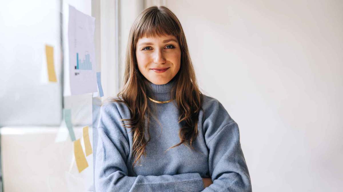 Braunhaarige Frau auf der Arbeit trägt einen hellblauen Pullover und eine goldene Kette. Sie steht mit verschränkten Armen und einem Lächeln vor einem Flipchart.