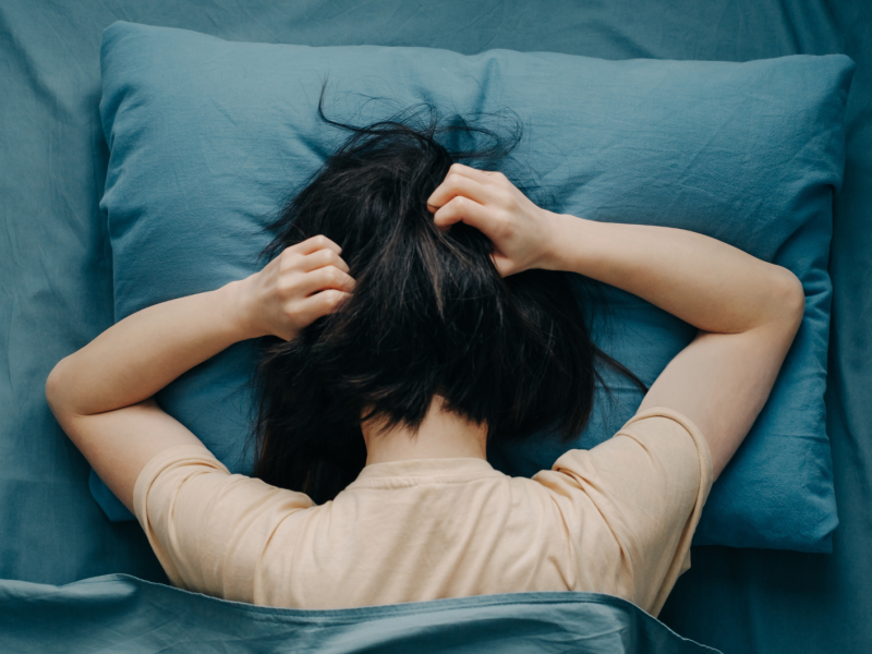 Eine junge Frau mit schwarzem Haar liegt auf einem blauen Kissen und greift sich mit beiden Händen in die Haare.