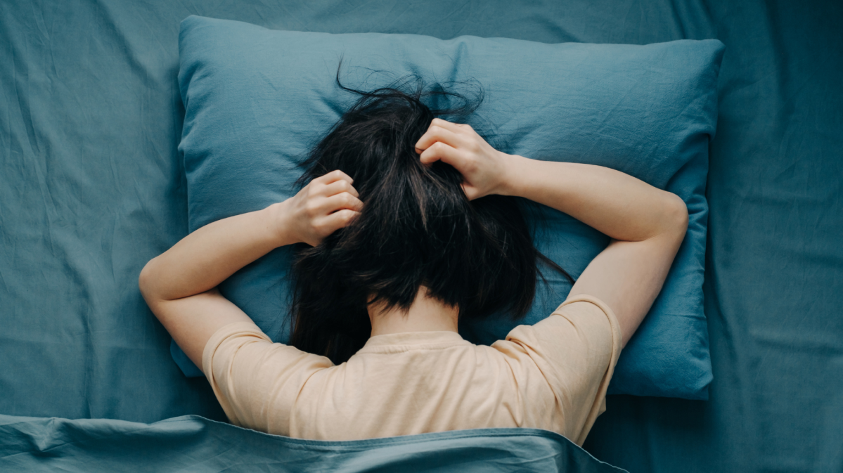 Eine junge Frau mit schwarzem Haar liegt auf einem blauen Kissen und greift sich mit beiden Händen in die Haare.