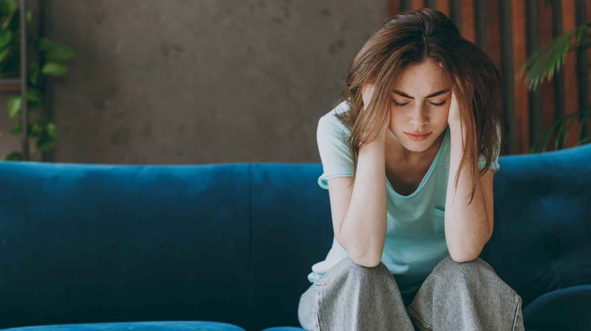 Frau sitzt auf einer Couch und guckt traurig zu Boden. Dabei hält sie ihren Kopf und greift in ihre Haare.