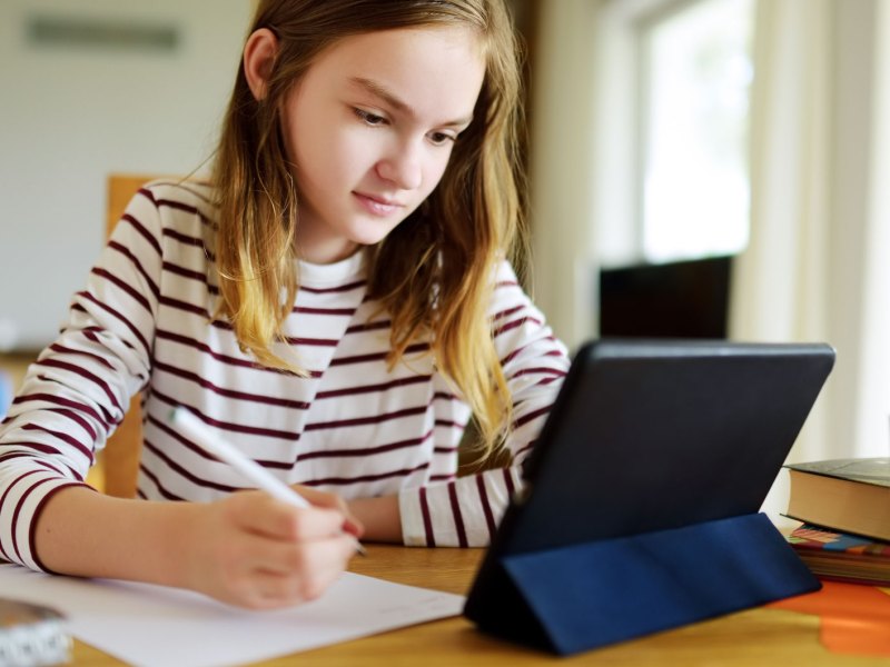 Junges Mädchen, ca. 12 Jahre, sitzt konzentriert am Schreibtisch vor einem Tablet und macht sich Notizen.