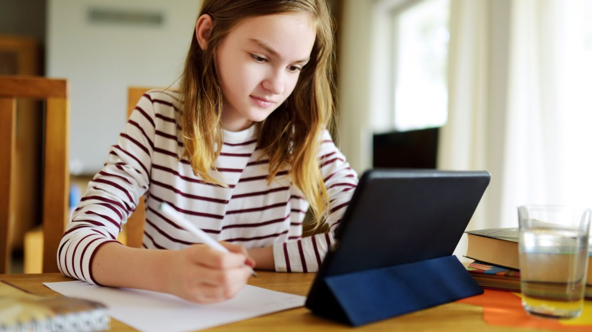 Junges Mädchen, ca. 12 Jahre, sitzt konzentriert am Schreibtisch vor einem Tablet und macht sich Notizen.