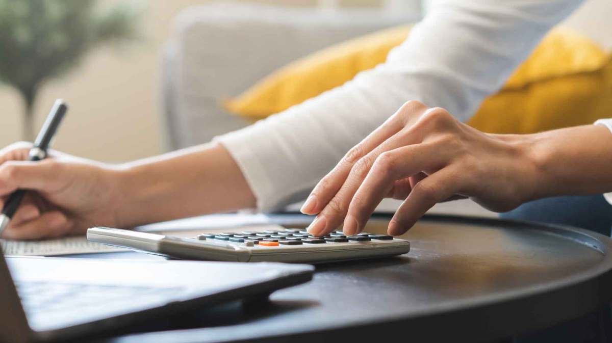 Frau sitzt zuhause an einem Tisch vor einem Laptop und rechnet mit einem Taschenrechner ihre Steuern aus.