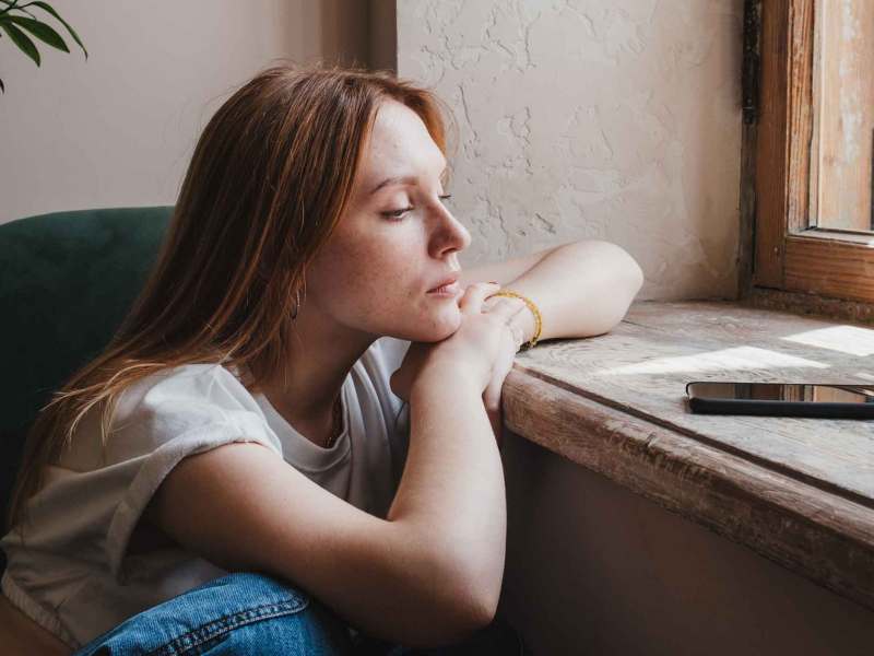 Frau sitzt am Fenster und guckt unglücklich auf ihr Handy, das auf der Fensterbank vor ihr liegt.