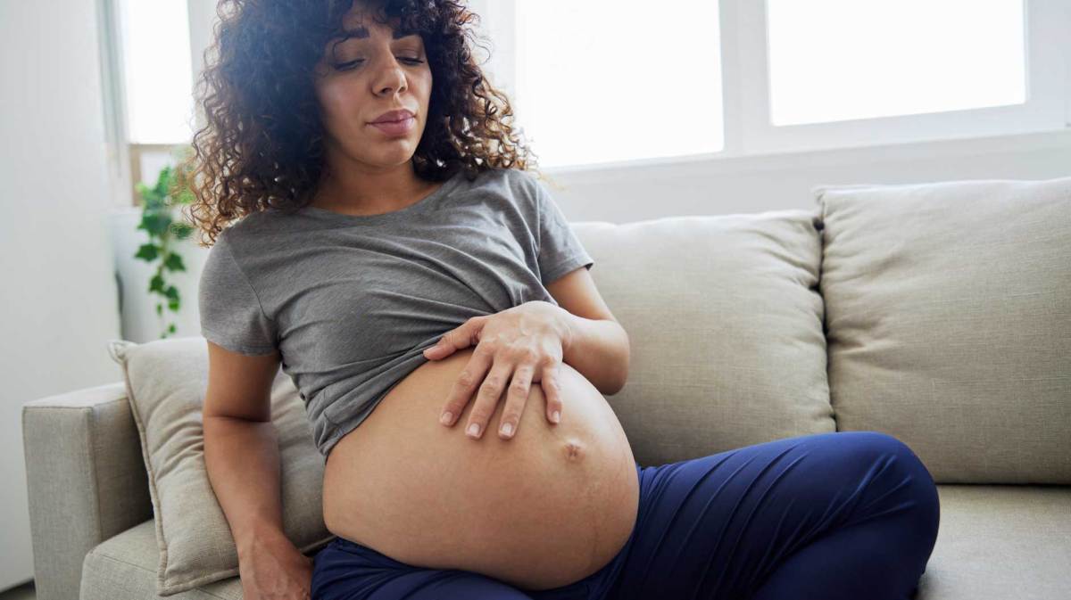 Schwangere Frau sitzt mit leicht schmerzverzerrtem Gesicht auf dem Sofa und hat eine Hand auf ihren Babybauch gelegt.