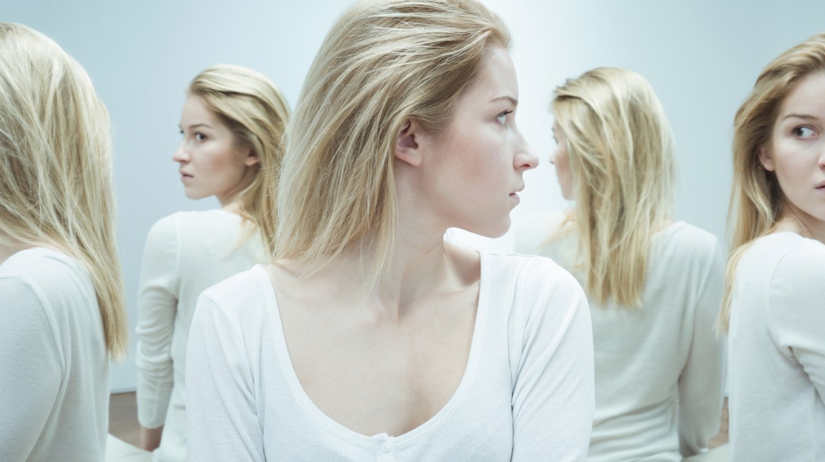 Blonde Frau sitzt vor mehreren Spiegeln und wird von Spiegelbildern angeschaut.