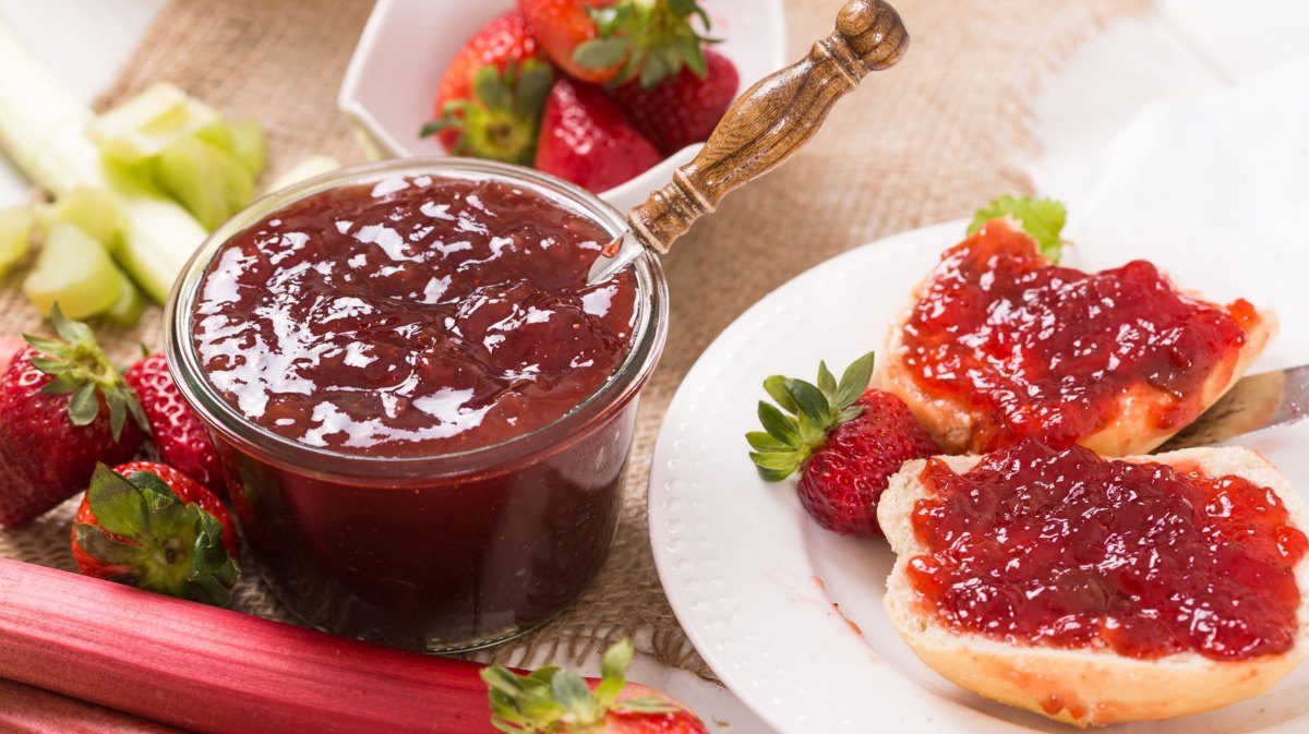 Fruchtaufstrich in Glas, neben weißem Teller mit zwei Brötchenhälften, die mit Marmelade beschmiert sind.