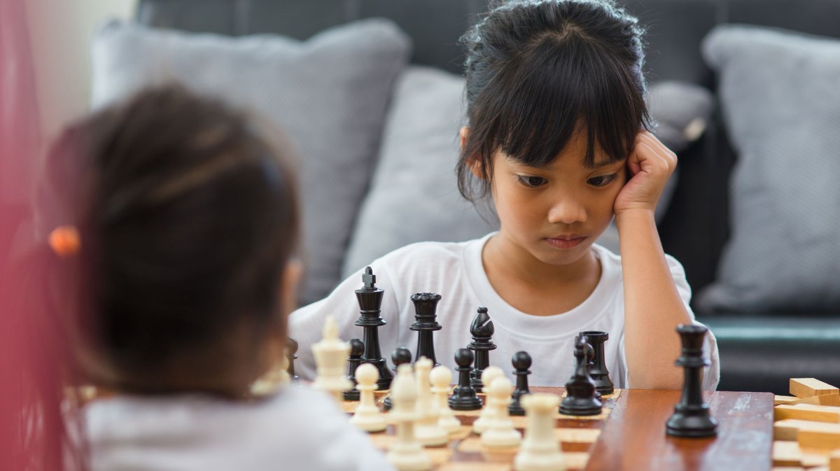 Zwei Mädchen, ca. 7 Jahre alt, spielen am Wohnzimmertisch eine Runde Schach. Ein Mädchen ist von hinten zu sehen, das andere von vorn. Es schaut konzentriert auf das Spielbrett.