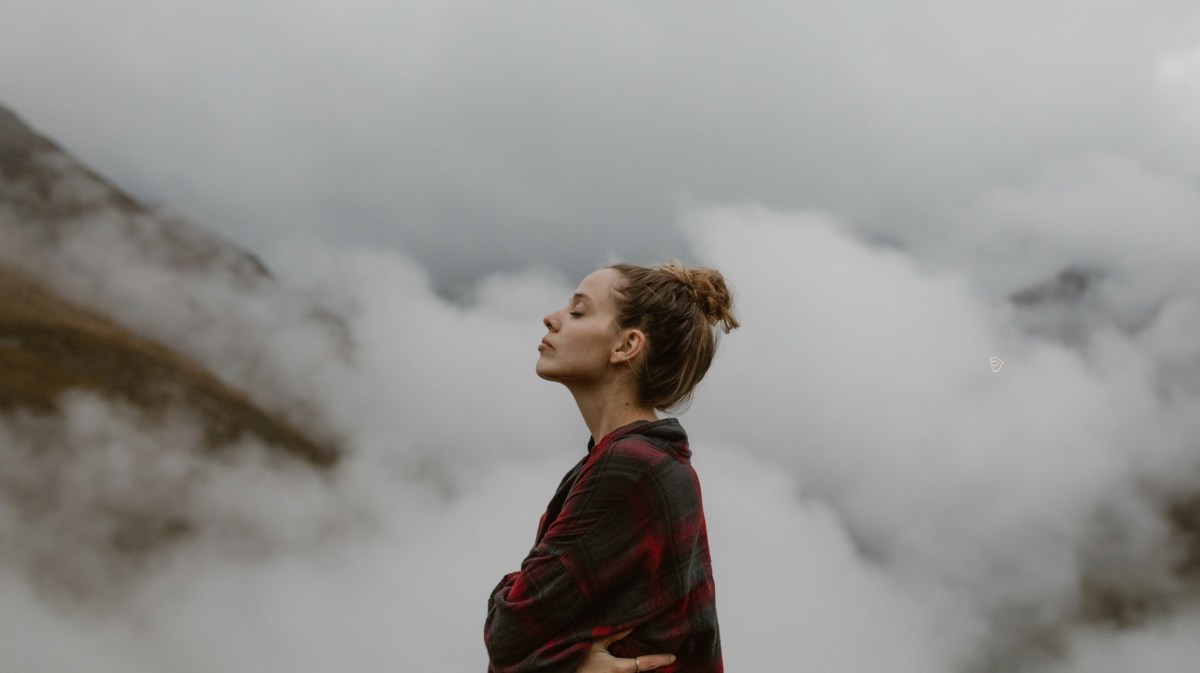 Frau auf einem Berg mit Wolken im Hintergrund, die ihre Augen schließt und einatmet