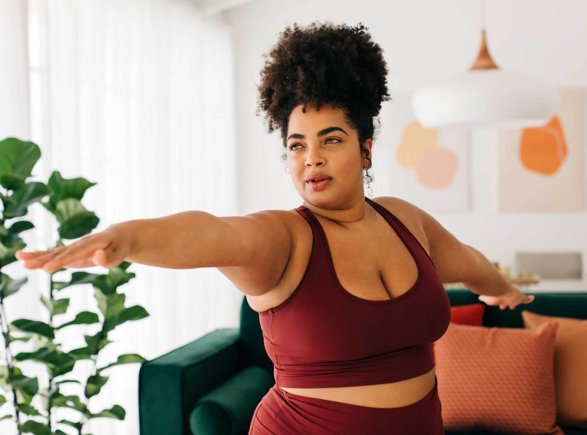 Junge Frau mit Afro und bordeauxrotem Sportoutfit balanciert beim Workout im hellen Wohnzimmer.