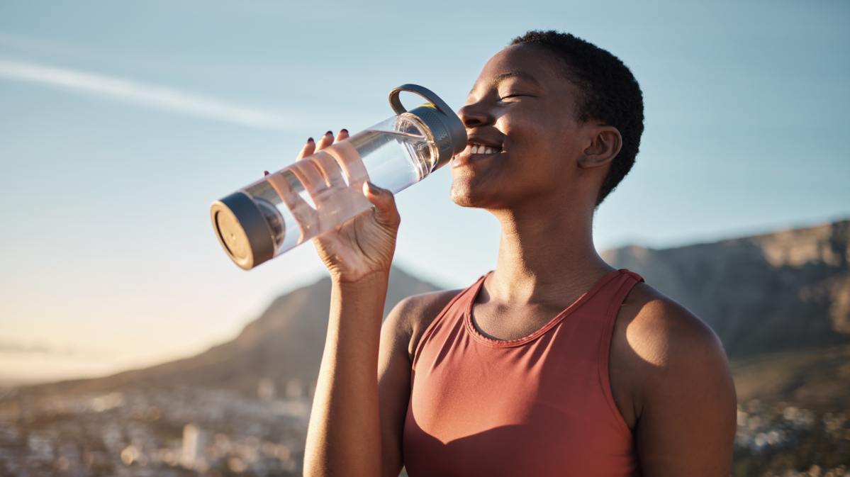 Frau auf einem Berg mit Landschaft im Hintergrund, die glücklich aus einer Wasserflasche trinkt.