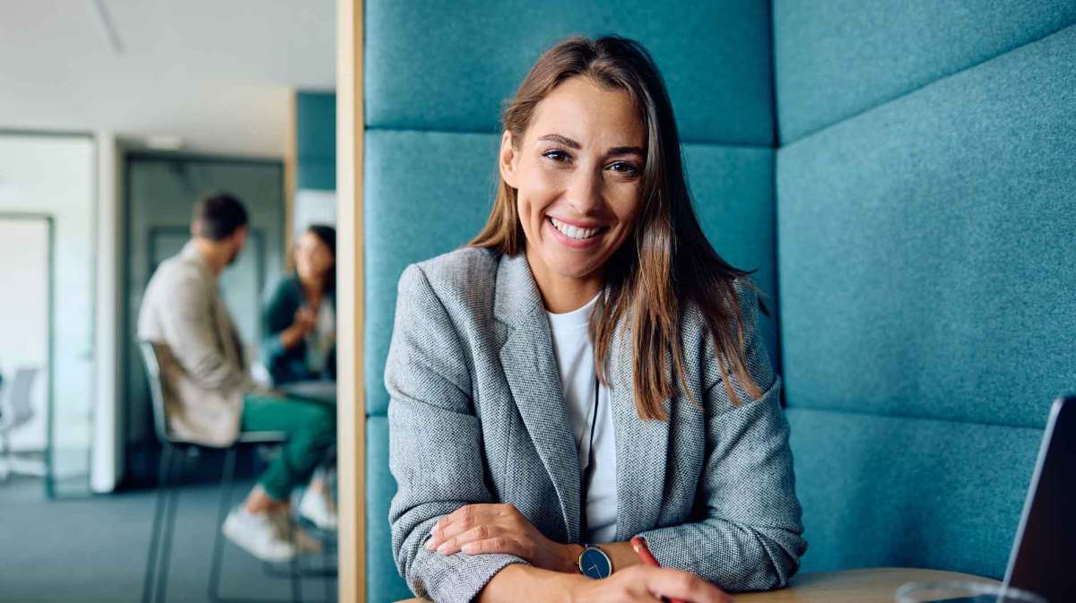 Junge Frau mit braunen Haaren und hellblauem Blazer sitzt im Büro und lächelt.