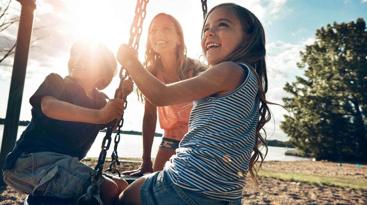 Mutter spielt mit Tochter und Sohn auf einem Spielplatz. Die Kinder sitzen auf einer Reifenschaukel.