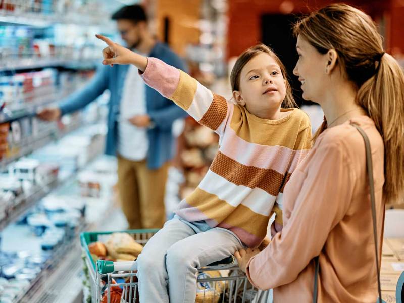 Mutter und Tochter stehen im Supermarkt vor einem Kühlregal. Die Tochter zeigt auf etwas, das sie haben möchte.