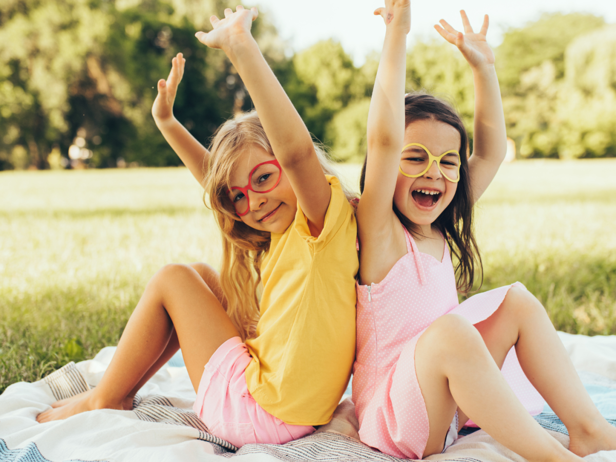 Zwei Kinder auf einer Picknickdecke, die Brillen anhaben und die Arme lachend in den Himmel strecken.