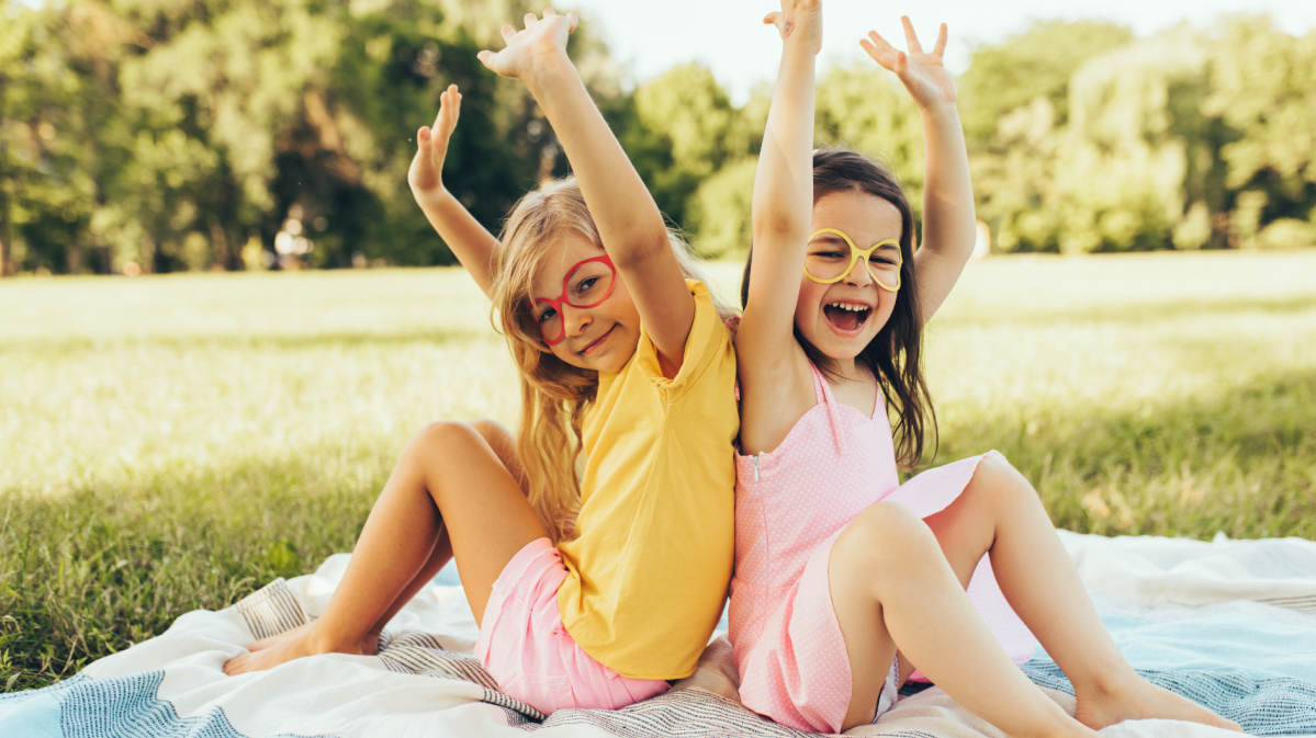 Zwei Kinder auf einer Picknickdecke, die Brillen anhaben und die Arme lachend in den Himmel strecken.