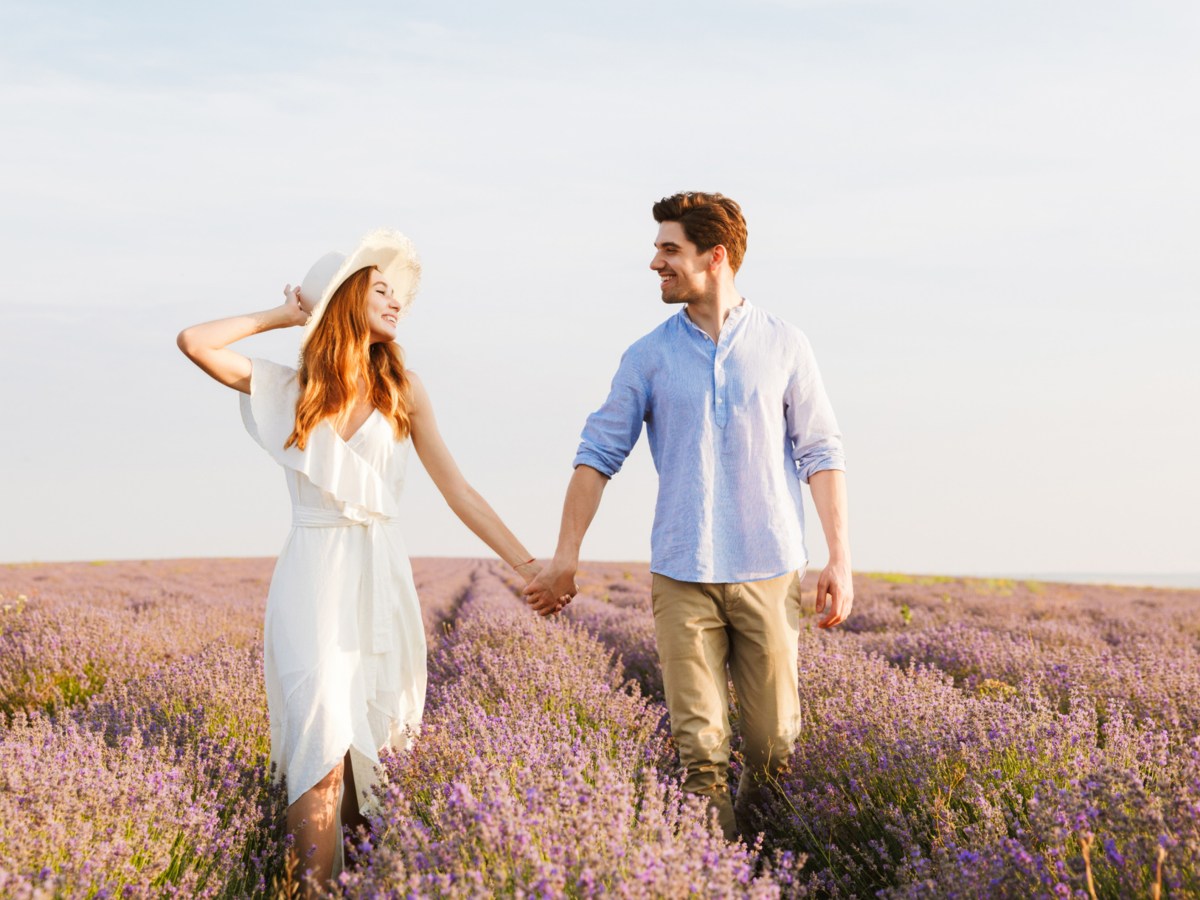 Frau und Mann die im Sommer auf einem Lavendelfeld Händchen halten und sich gegenseitig anlächeln.