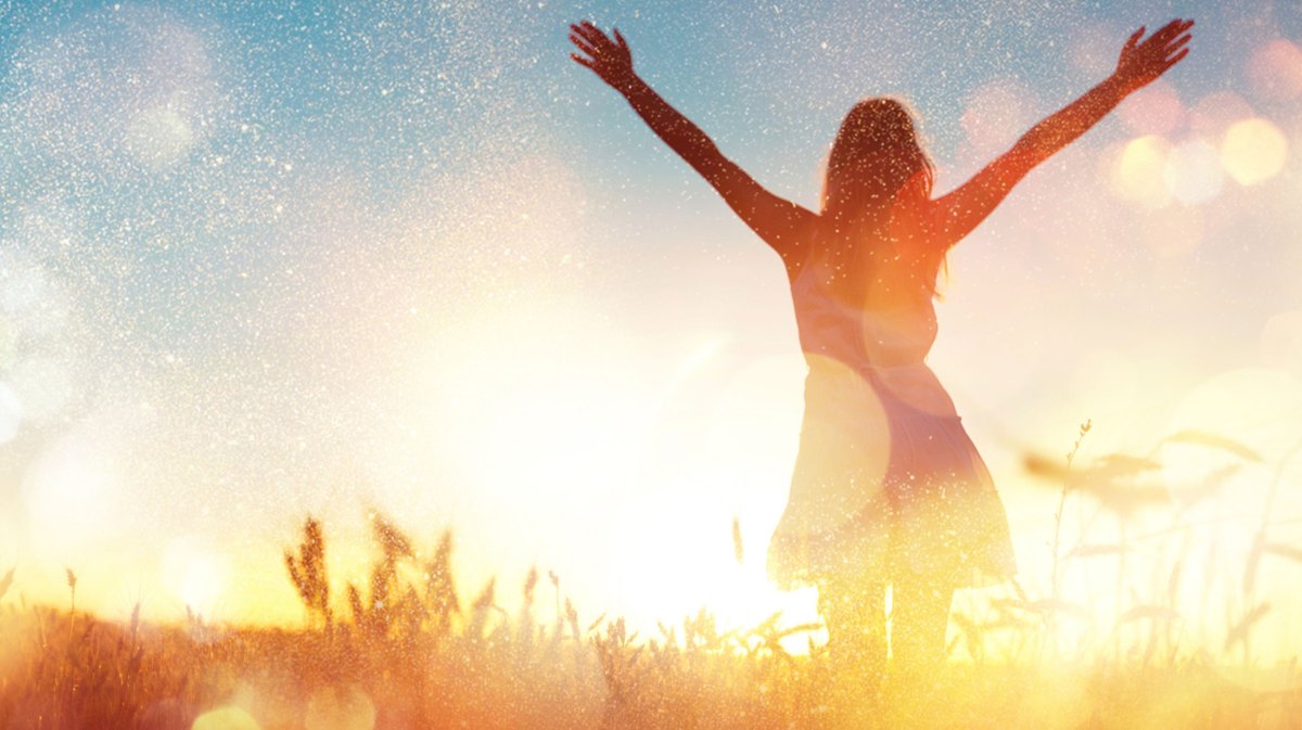 Frau im Sommerkleid mit ausgestreckten Armen auf einem Feld bei Sonnenuntergang