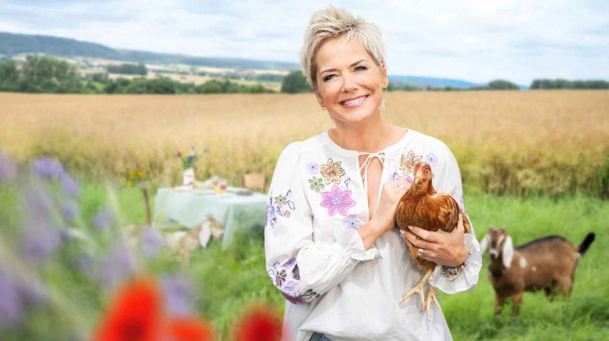 Inka Bause steht auf einem Feld und hält ein Huhn im Arm.