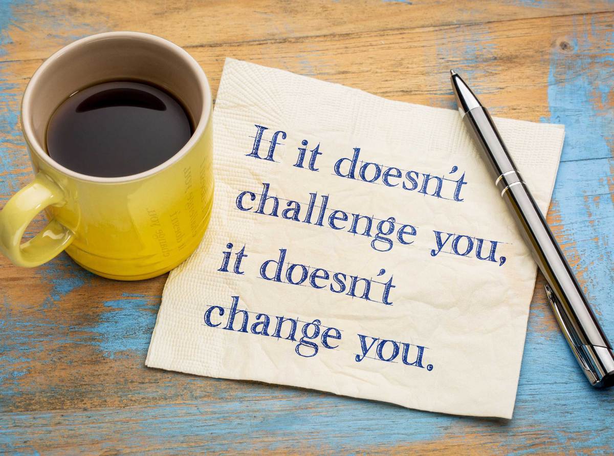 Gelbe Kaffeetasse neben einer Serviette auf einem Holztisch. Notitz auf Serviette: "If ist doen't challenge you, it doesn't change you."