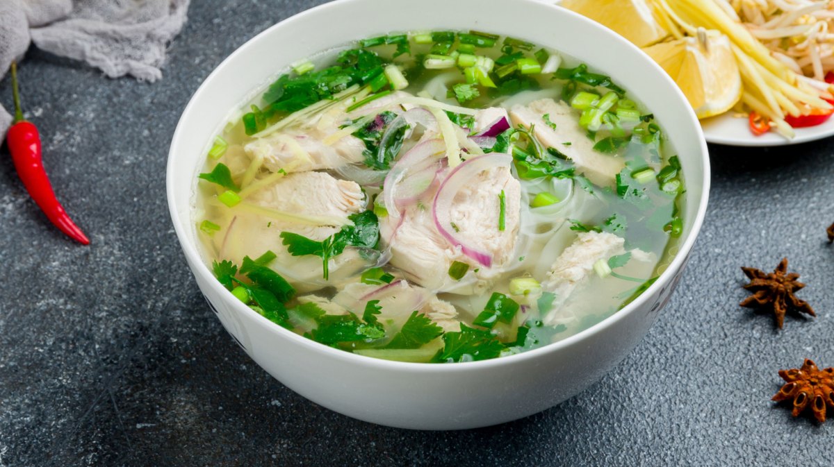 Pho ga Suppe in einer weißen Schüssel mit Zutaten, die da rum verstreut wurden.