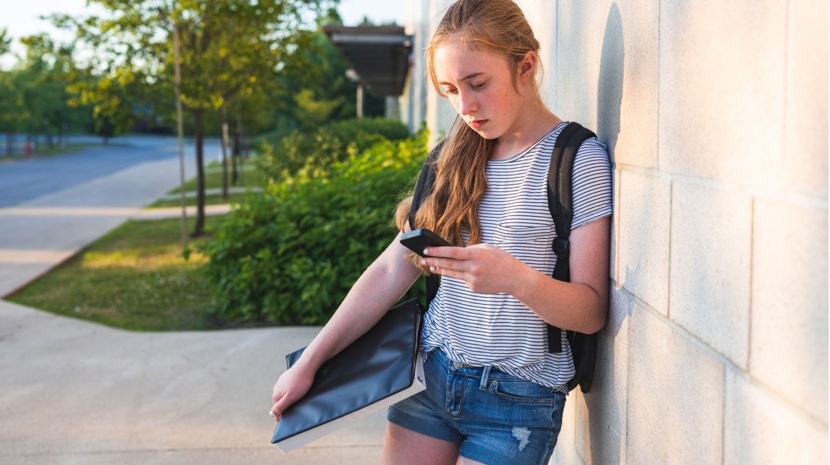 Mädchen in Shorts und Shirt, mit Büchern unterm Arm, steht an die Wand gelehnt und schaut traurig auf ihr Handy.