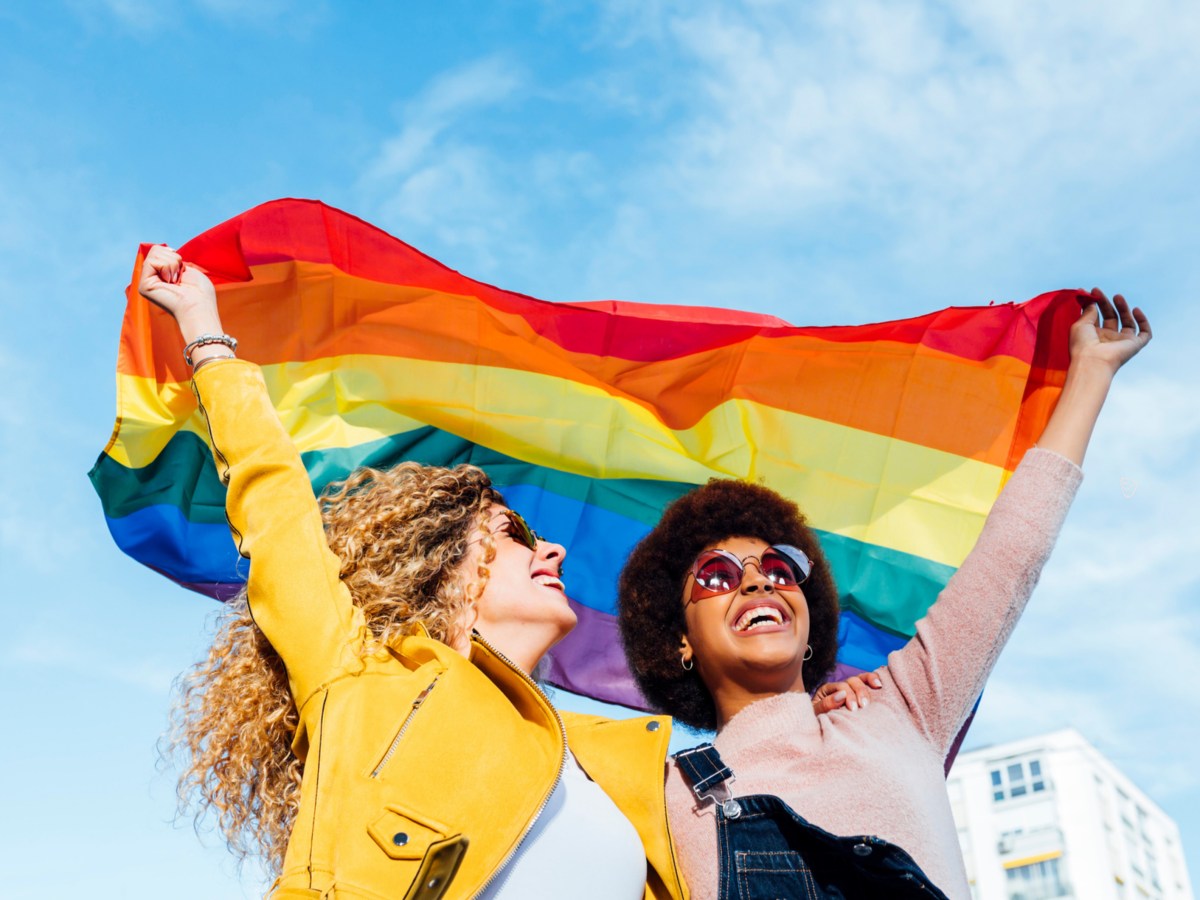 Zwei Freund*innen, die eine Pride-Flagge hochhalten und glücklich nach vorne blicken.