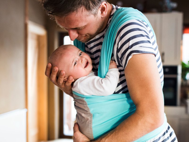 Vater hat sein Baby in einem Tragetuch vor dem Bauch. Das Kind ist unglücklich mit der Situation.