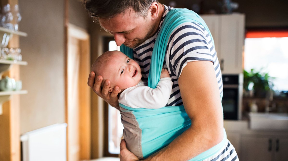 Vater hat sein Baby in einem Tragetuch vor dem Bauch. Das Kind ist unglücklich mit der Situation.